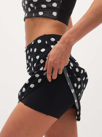 Model in action wearing Club Skort in Black with white polka dots, featuring a built-in short and bow detail.