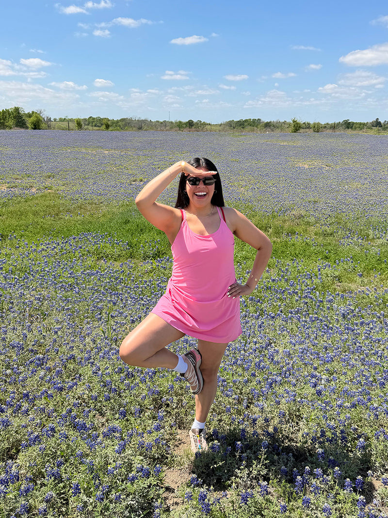Model posing in The Exercise Dress in Pink with adjustable straps and built-in shorts liner, set in a field of bluebonnets.
