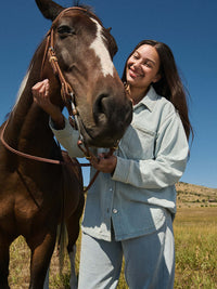 Smiling model with a horse wearing Desert Pant in light blue, wide-leg denim cotton terry sweatpant with sun-scorched effect.
