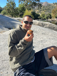 Person sitting by a river wearing RecFleece 1/2 Zip in Olive, enjoying a muffin in a relaxed outdoor setting.