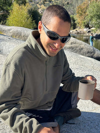 Man sitting on a rock wearing RecFleece 1/2 Zip in Olive, holding a coffee cup while enjoying nature.