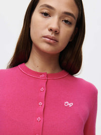 Model posing in Sugar Cardigan in bright pink, featuring hand-embroidered OV signature and button details.