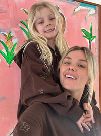 Two models wearing Sunny Hoodies in Brown, featuring playful designs on sleeves, smiling against a colorful background.