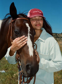 Woman wearing a Doing Things Trucker Hat, affectionately posing with a horse against a scenic backdrop.