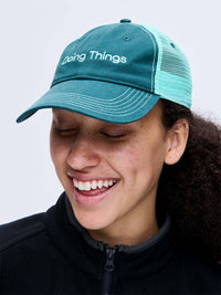 Close-up of smiling model wearing Trucker Hat in Teal, featuring breathable mesh back and embroidered Doing Things logo.
