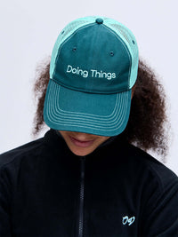 Close-up of model wearing Trucker Hat in Teal, featuring 'Doing Things' embroidered on the front.